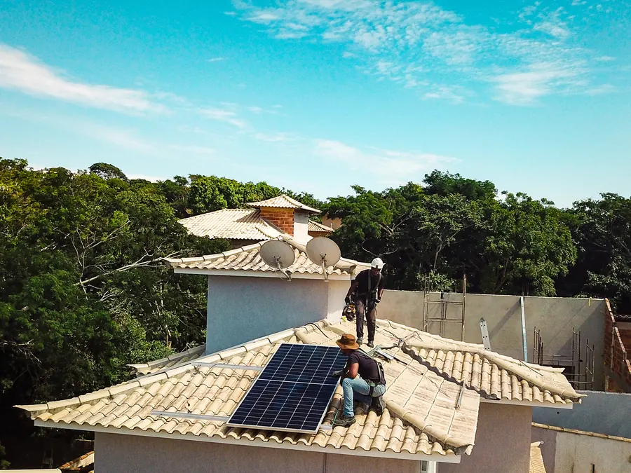 Equipe instalando módulos em casa com telhado cerâmico, com mata ao fundo